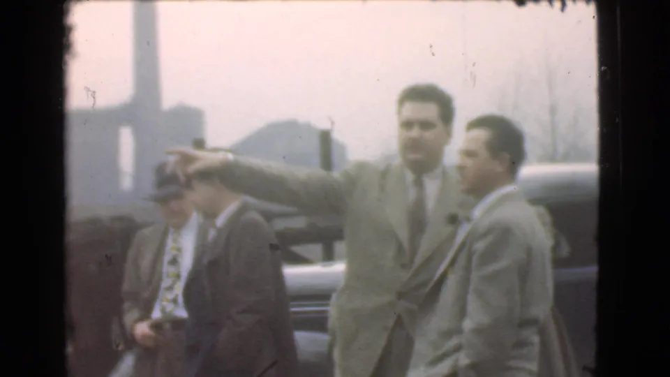 In the Cleveland home-movie frame, the central man points left while men in hats and suits stand beside a dark automobile.