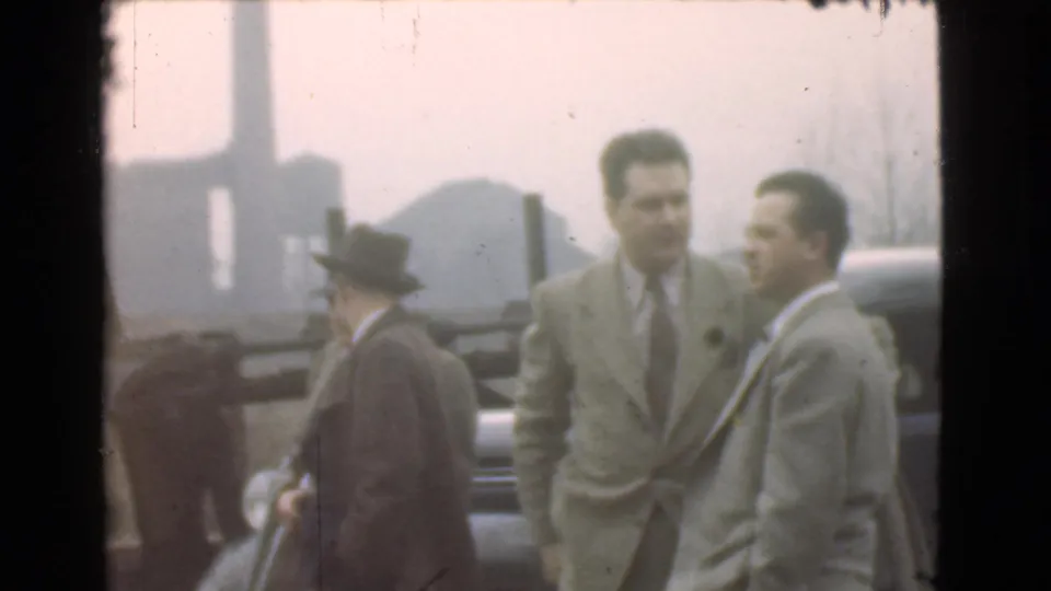 The group of adult men remains beside a parked car in 1939 Cleveland, with formal coats and hats visible in soft 8mm color.