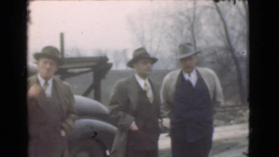 Three adult men in hats and coats stand near an automobile in the Cleveland outdoor scene, with bare trees and muted winter light behind them.