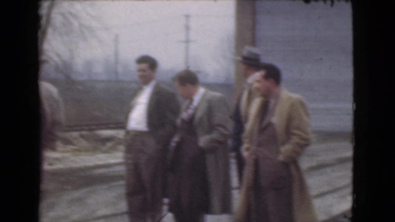 Five adult men in coats and suits stand near a street with utility poles in the background.