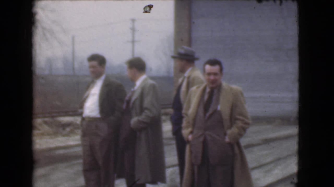 A small group of men in overcoats stands on a street, with one man facing the camera.