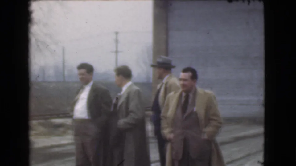 Adult men in 1939 Cleveland street footage stand casually in long coats, with a hat and utility poles visible behind them.
