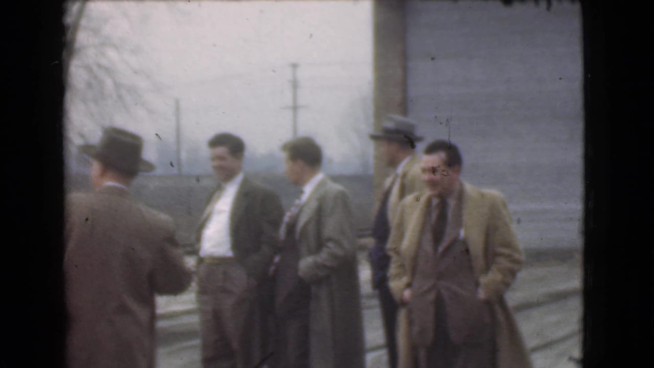 Five adult men in suits and coats stand outdoors near a building and utility poles.