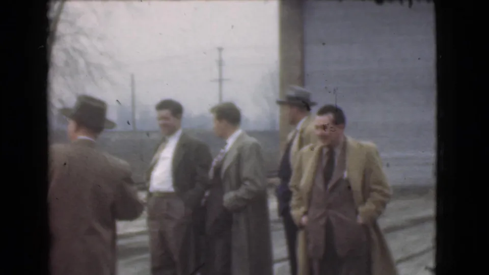 Five adult men in 1939 Cleveland stand outdoors in suits, overcoats, and hats near a building wall and utility poles.