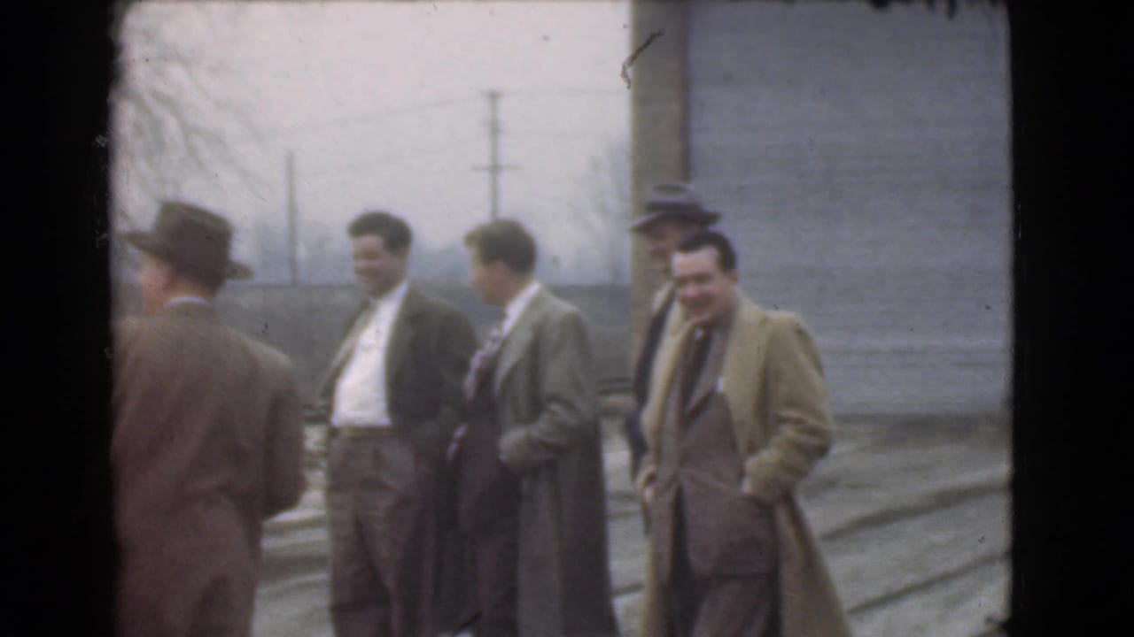 A group of men in coats and hats converse outdoors beside a building.