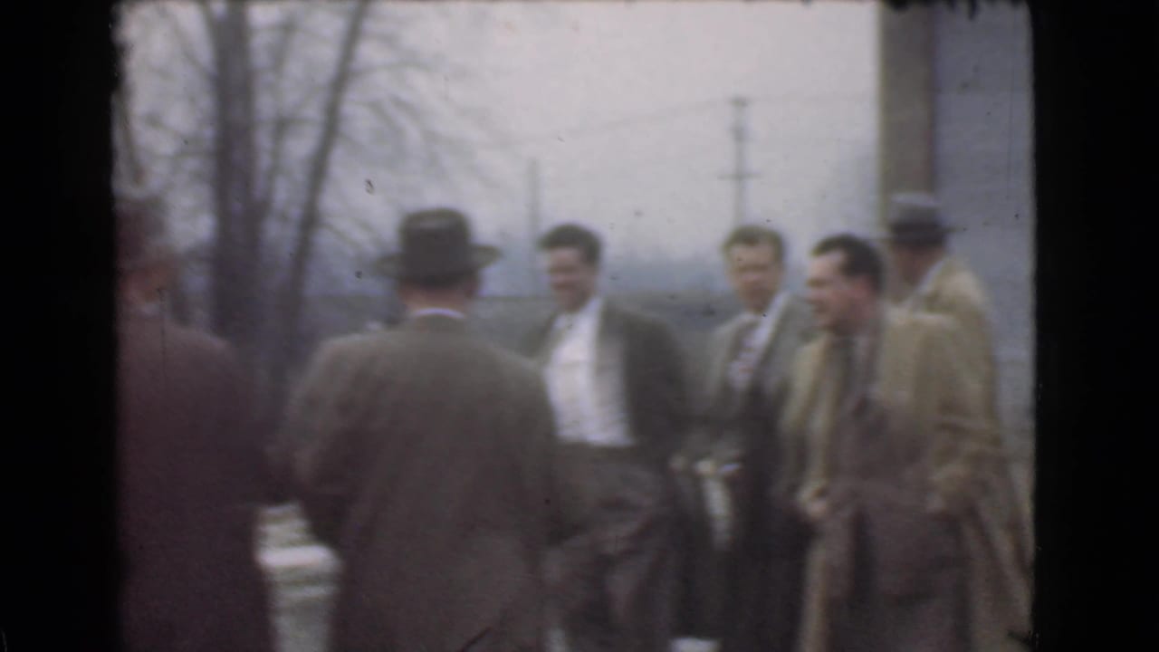 Several men in suits and hats stand outdoors near bare trees and utility poles.