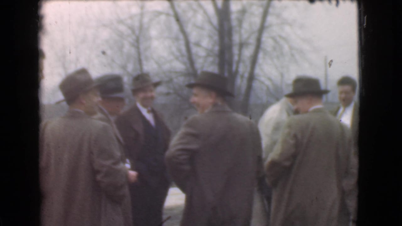 A group of men in hats and overcoats converse outdoors beside leafless trees.
