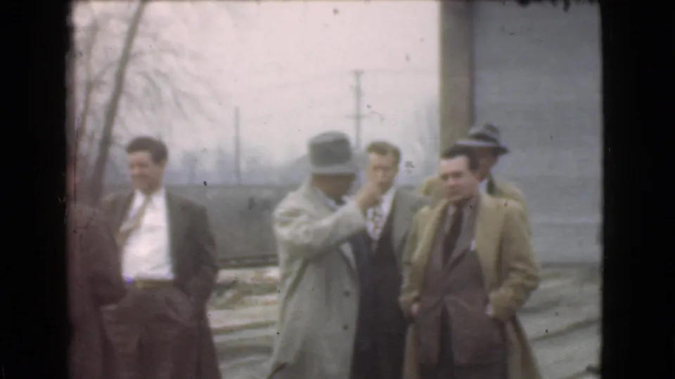 Adult men in fedoras and overcoats remain grouped beside the street in Cleveland, Ohio, 1939 home-movie footage.
