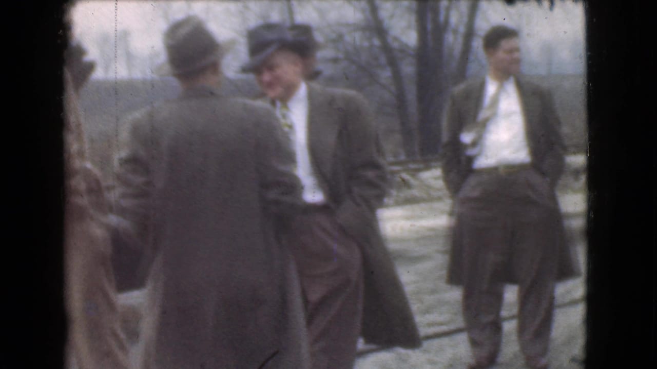 Men in suits and hats stand outdoors near a road and bare trees.