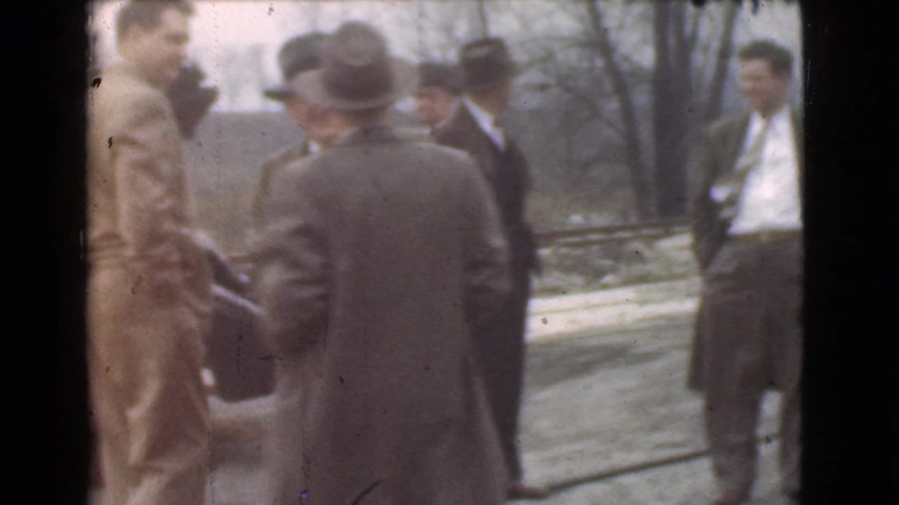 Adult men in suits and hats appear to converse outdoors in Cleveland, with a car partially visible at the side of the frame.