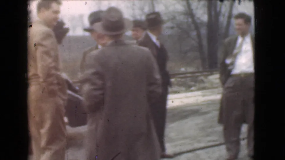 Adult men in suits and hats appear to converse outdoors in Cleveland, with a car partially visible at the side of the frame.