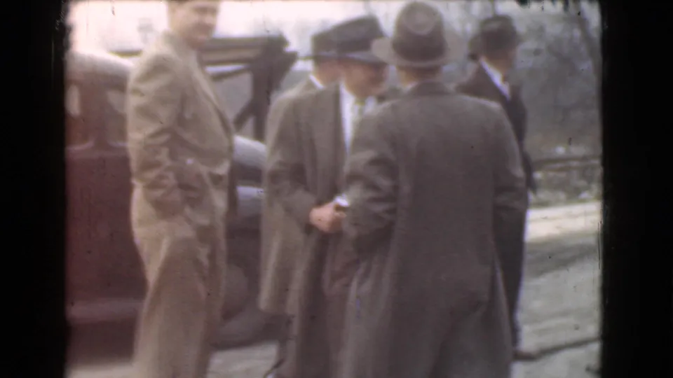 The home-movie frame shows suited men in hats gathered near a parked automobile, with bare trees and a roadside behind them.