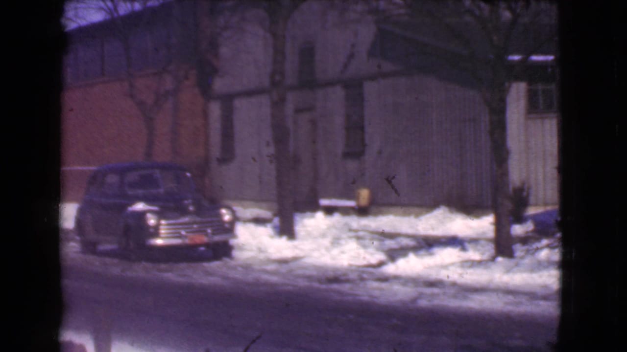Dark vintage car parked on a snowy street beside bare trees and a brick building.