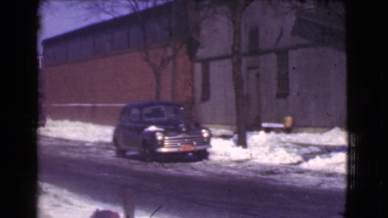 Parked dark car on a slushy street with snow piled near a brick-and-gray building.