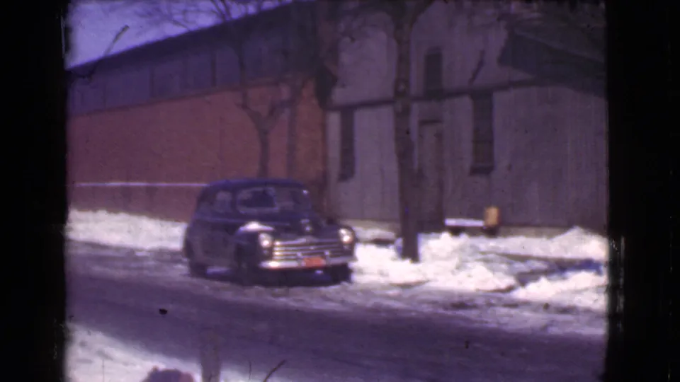 1939 Cleveland winter street view showing the parked car closer to center, with snowbanks, bare trees, brick wall, and gray siding in soft 8mm focus.