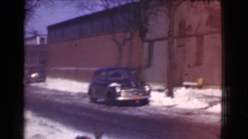 1939 Cleveland home-movie frame showing a parked dark automobile on a slushy winter street, with a brick building, bare tree, snowbanks, and part of another car at left.