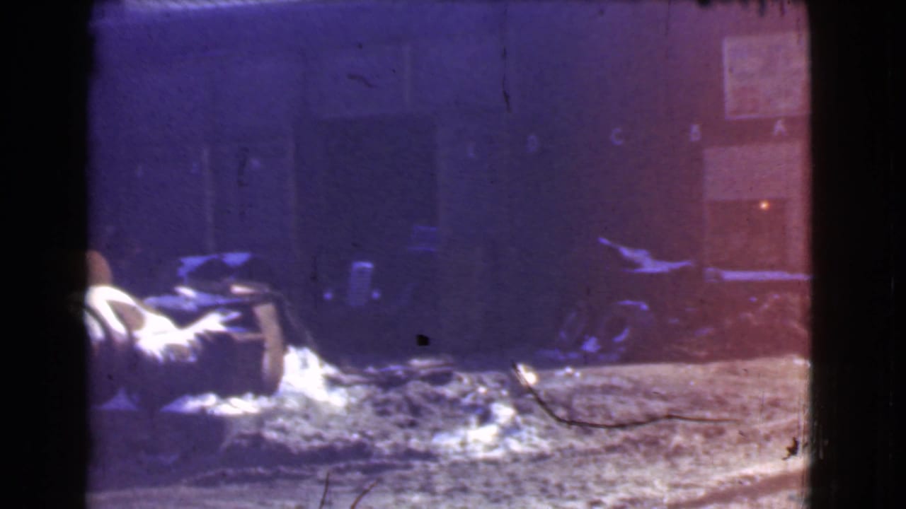 Dim industrial yard with barrels, debris, snow patches, and a dark warehouse wall.