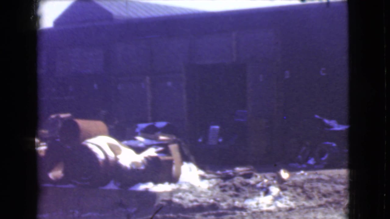 1939 Cleveland home-movie frame with large drums or barrels on rough ground outside a dark industrial building.