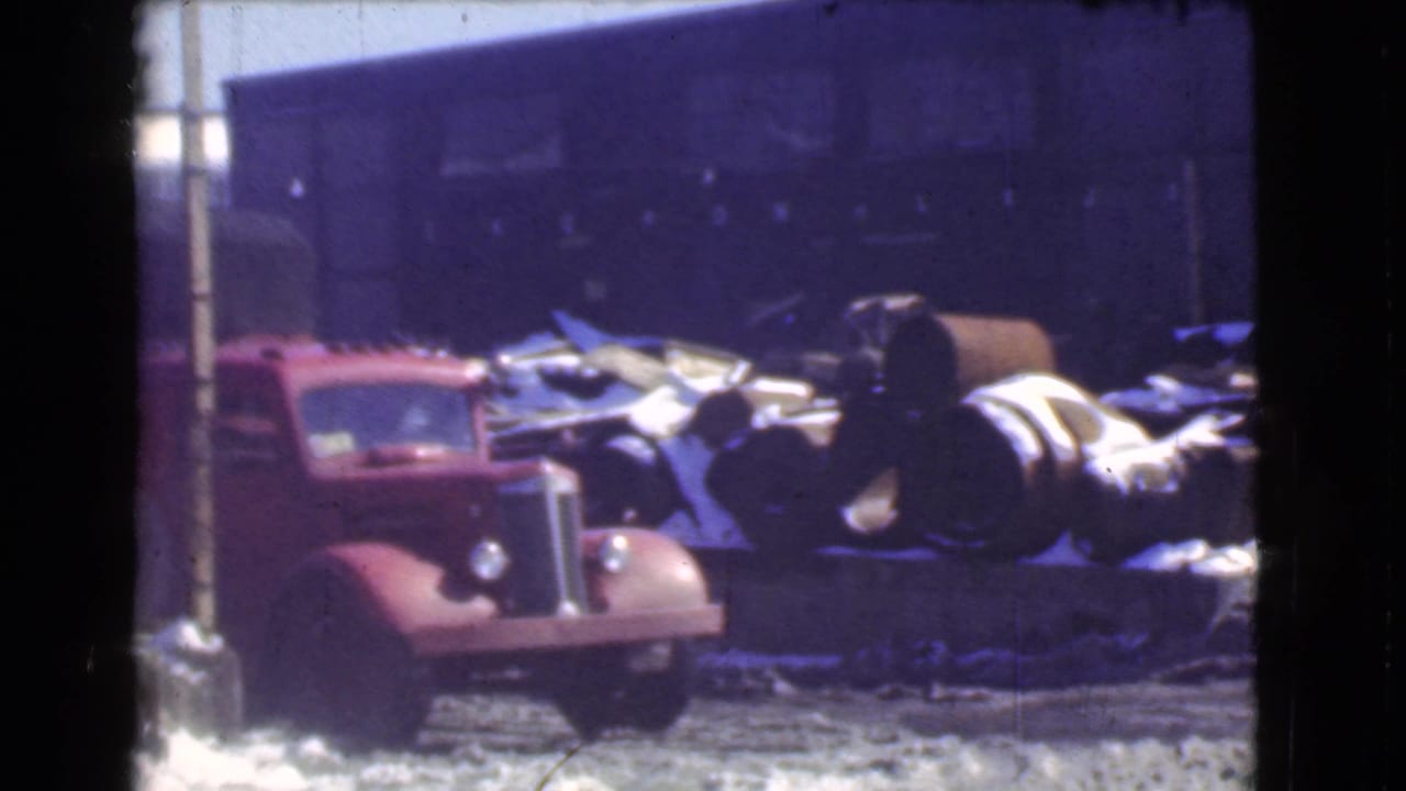 Front of a red truck parked beside barrels outside a factory building.