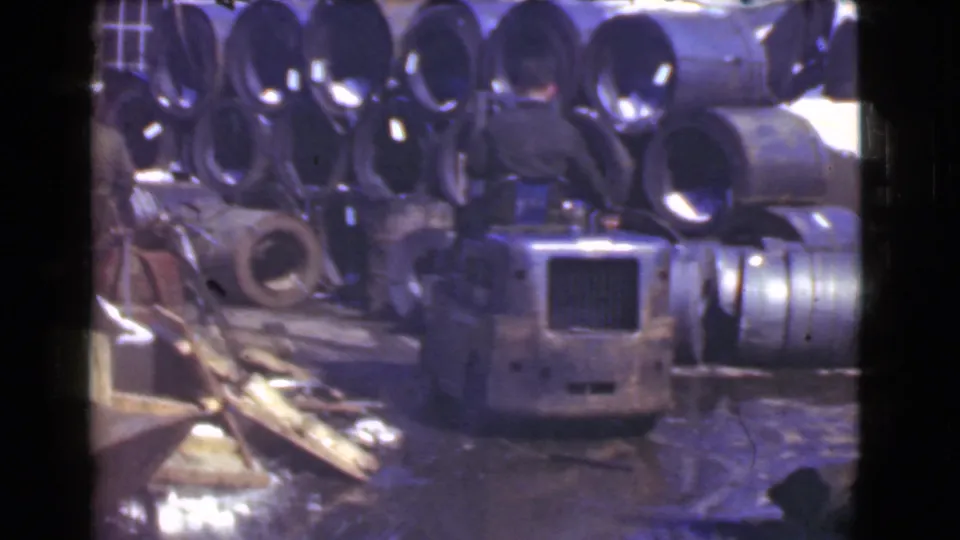 A worker stands by a forklift-like vehicle in a Cleveland scrap yard with large pipes stacked behind him.
