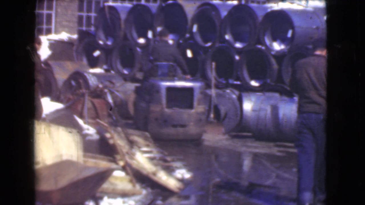 Workers near stacked metal pipes and machinery in a wet factory yard.