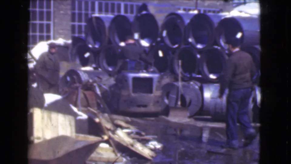 Two workers in a Cleveland factory yard, 1939, standing among stacked metal pipes, equipment, and scattered industrial materials.