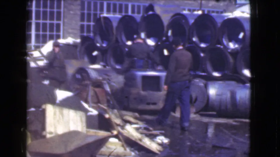 A worker walks through a Cleveland factory yard in 1939, passing stacked metal pipes, machinery, and wet ground.
