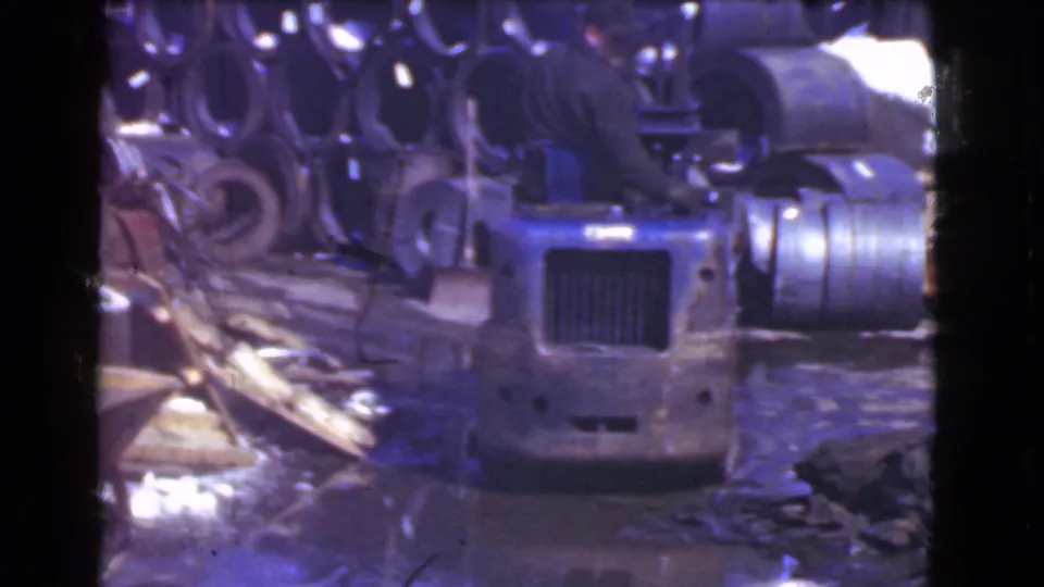 A small forklift faces the camera in 1939 Cleveland footage, with stacked metal cylinders and a wet debris-strewn floor around it.