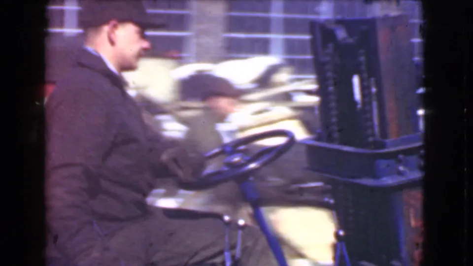 A capped worker drives the blue forklift through a Cleveland industrial area, with a brick wall and blurred vehicle in the background.