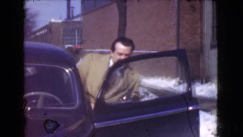 In Cleveland, a man in a light winter coat stands beside an open black car door near brick industrial buildings and snow.