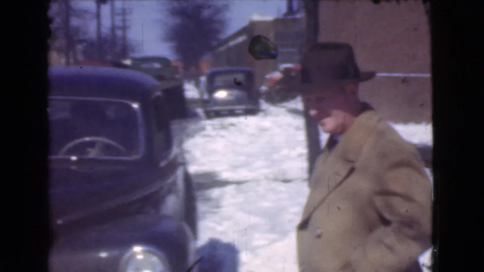 A man in a brown overcoat and brimmed hat walks along a snowy Cleveland industrial street beside dark parked cars.