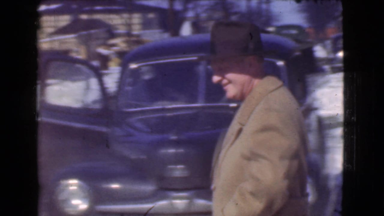 In Cleveland, Ohio, a man in a fedora-like hat and tan winter coat walks past rounded parked cars on a snowy 1939 street.