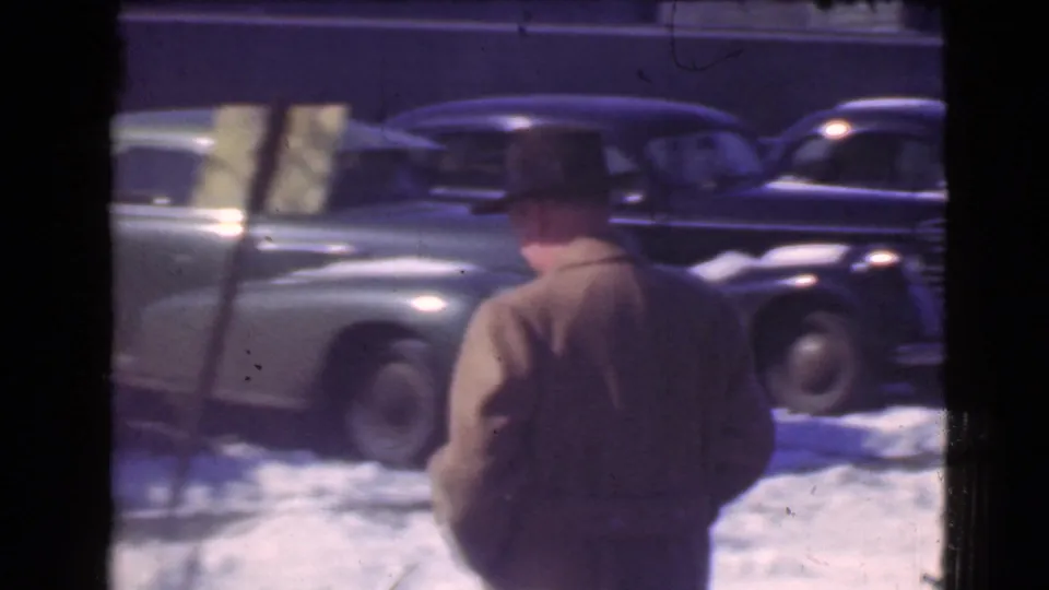 Rear view of the hat-wearing man walking past snow and passing automobiles on a Cleveland winter street in 1939.