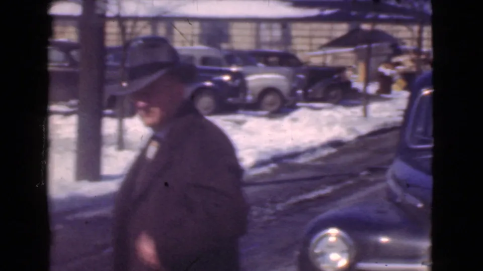 A hat-wearing man in a dark winter coat is framed near a snowy curb with parked 1930s cars in Cleveland, Ohio.