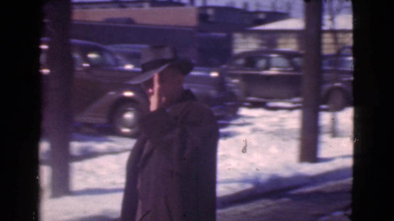 Man in a dark coat and fedora walking beside snowy parked cars.