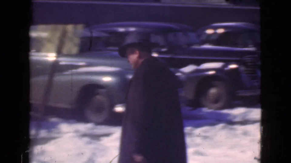 The man continues along a snowy Cleveland street, with rounded 1930s sedans parked and passing behind him.