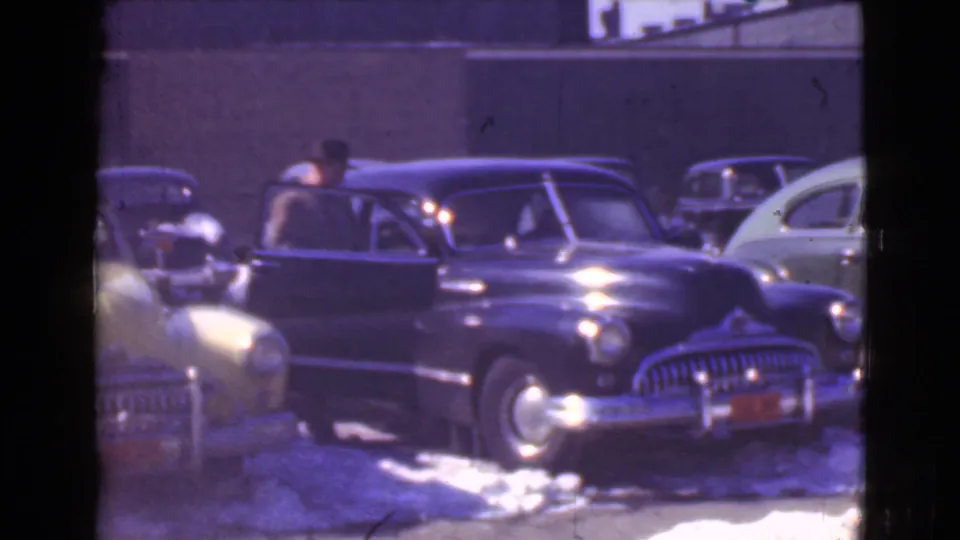 The man stands at an open door of a dark sedan on a snowy Cleveland street, surrounded by other period cars.