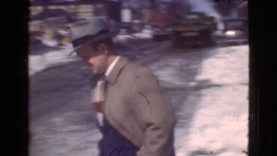 A man in a light coat and brimmed hat walks along a snowy Cleveland street with period cars behind him.