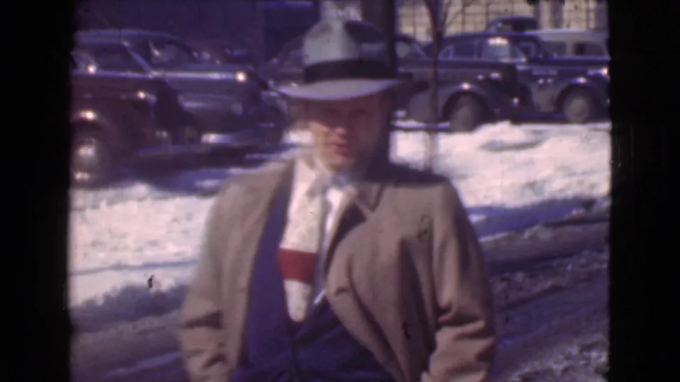 A suited man in a coat and brimmed hat faces the camera on a snowy Cleveland street with parked 1930s cars behind him.