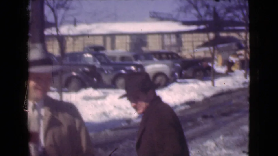 Two men in winter coats and hats appear near a snowy Cleveland curb with several period automobiles and a low building behind them.