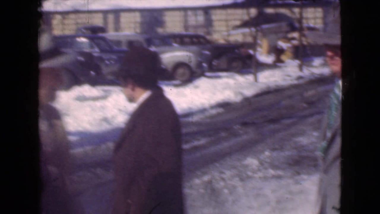In 1939 Cleveland, a man in a dark hat and overcoat moves along a snowy street lined with parked prewar cars.