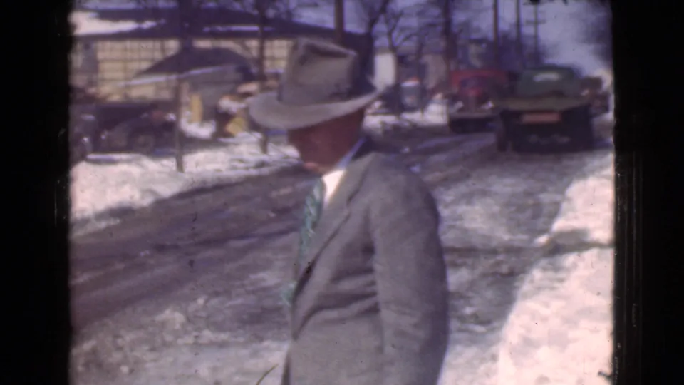 A fedora-wearing man in a suit is seen near a snow-covered Cleveland street with several period cars behind him.