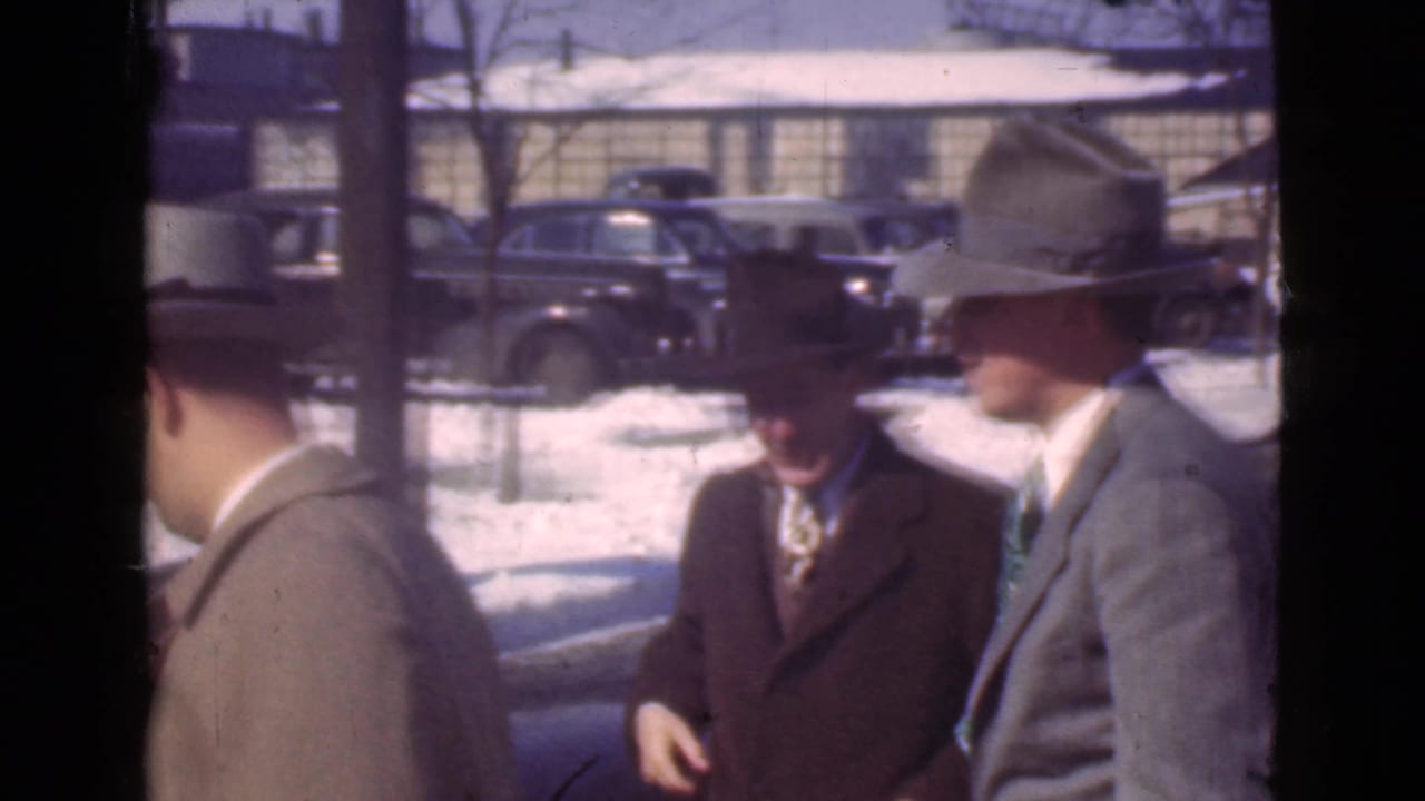 Three well-dressed adult men in suits, overcoats, and hats stand near vintage cars in snowy Cleveland, Ohio, footage from 1939.