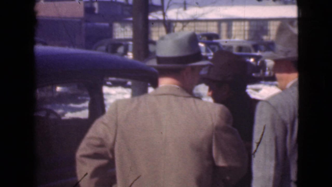 Men in hats are seen from behind beside a dark car in a snowy parking area.
