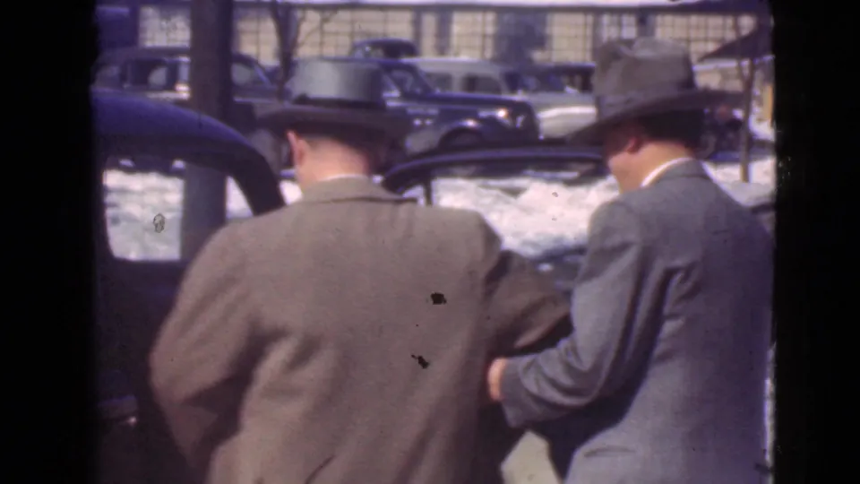 Two suited men in hats are filmed from behind near a dark car, with snow and several vintage automobiles visible in the Cleveland street setting.