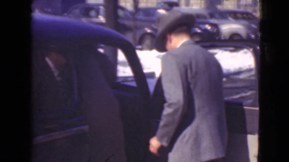 A suited man in a fedora-style hat moves toward or into a dark automobile with its door open, with snowy ground and parked cars behind him in 1939 Cleveland footage.