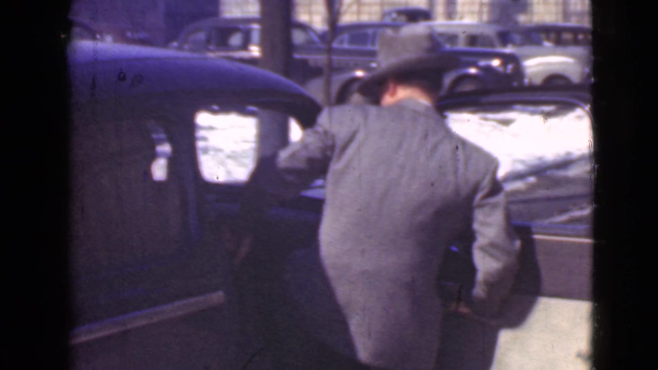 In 1939 Cleveland home-movie footage, a man in a suit and fedora stands beside a dark parked car on snow-covered pavement.
