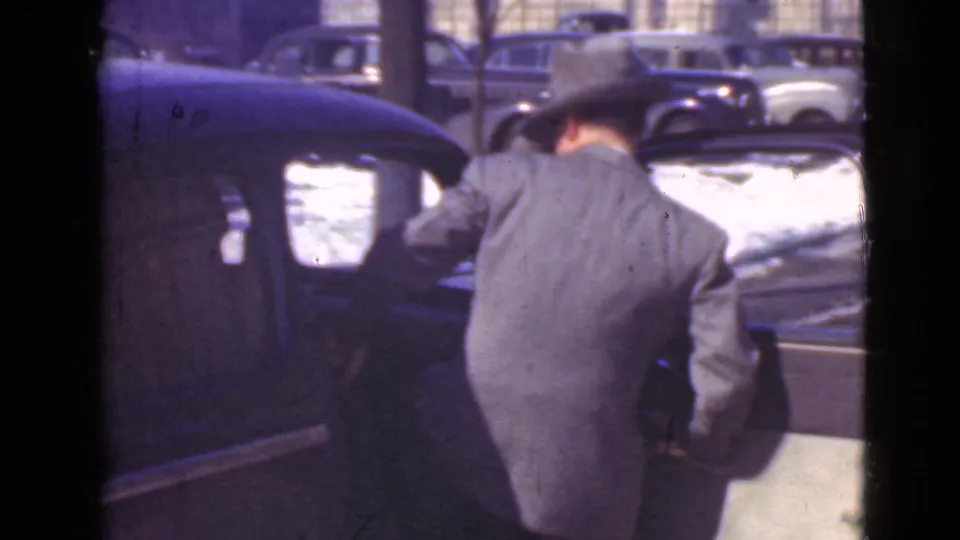 In 1939 Cleveland home-movie footage, a man in a suit and fedora stands beside a dark parked car on snow-covered pavement.