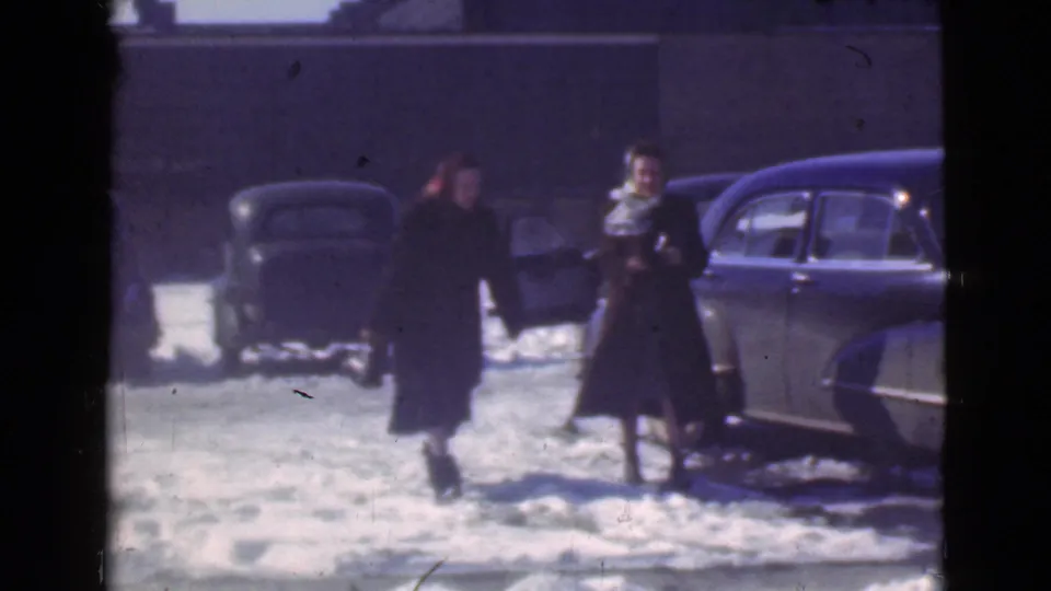 Two women in long winter coats walk through a snowy Cleveland parking lot, with 1930s cars and a building wall behind them.