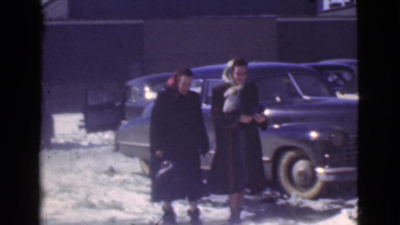 Two women in dark winter coats walk through a snowy parking lot near parked cars.